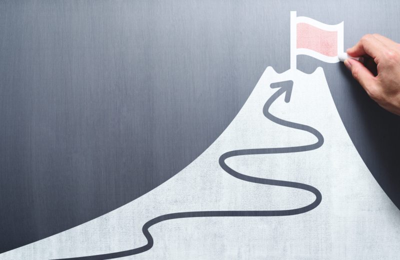 Business goal and success concept. Businessman drawing flag and mountain on chalkboard.